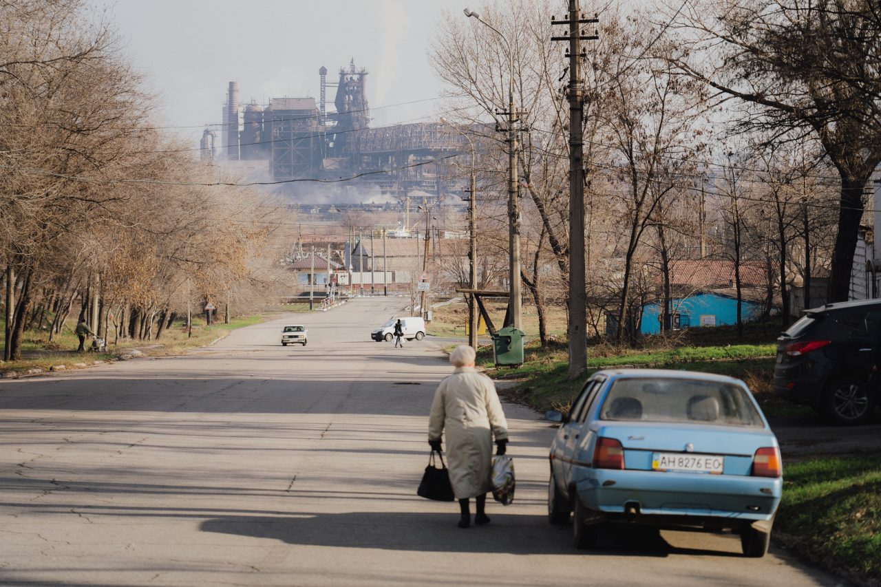 stará paní s taškami v rukou jde po silnici o kolo modrého auta, v pozadí fotky je kovový tovární kolos