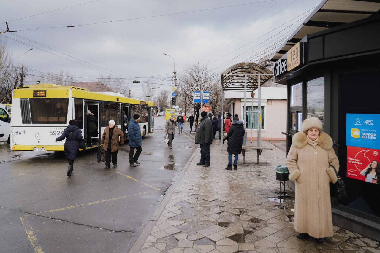 lidé čekající na zastávce na autobus, v popředí usměvající se paní 